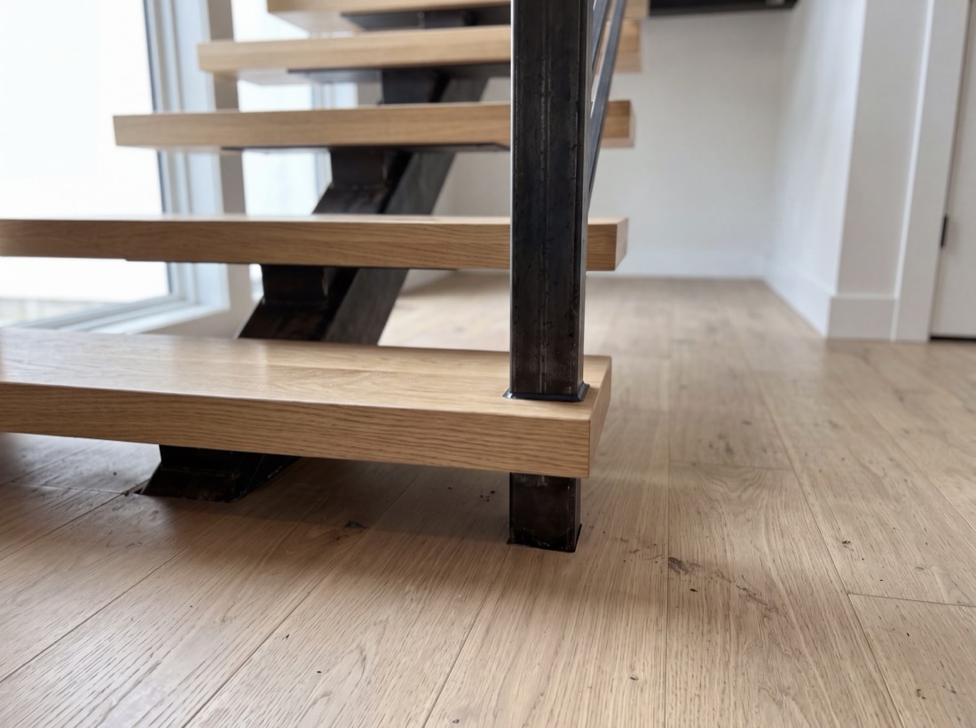 The base of an open-riser staircase, white oak treads meeting a blackened steel stringer